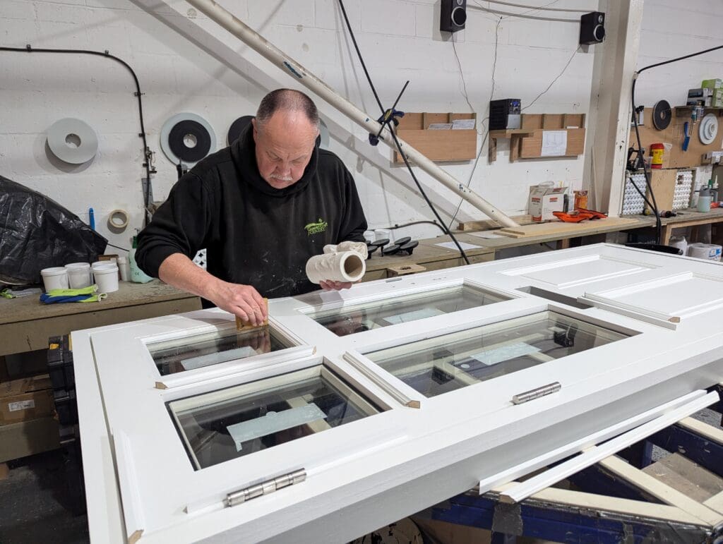 A skilled craftsman at Gowercroft Joinery carefully applies the final touches to a white timber door in the workshop. The high-quality glazing is securely fitted into the frame, ensuring a precise and weatherproof seal. The background features neatly organised tools, materials, and workstations, showcasing the attention to detail and expertise in traditional joinery.