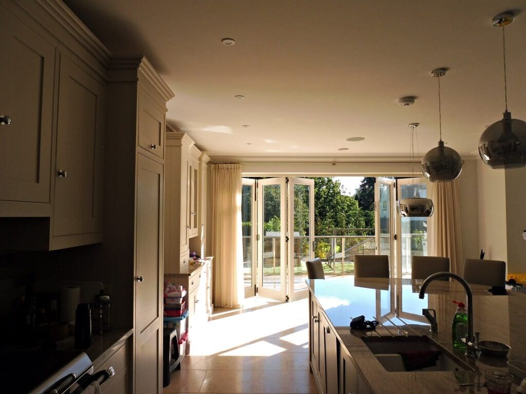 A five-panel Tutbury bifold door in painted Strutt Yellow, partially open, flooding a classic kitchen with natural light. The clear glazed panels create a seamless connection to the outdoor space, while the slim timber frames maintain a refined, traditional aesthetic. Perfect for blending indoor and outdoor living, this timber bifold door enhances both modern and period homes with its smooth operation and elegant design.