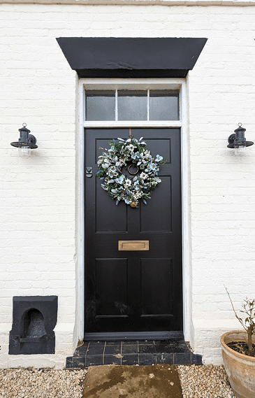 The entrance of a white brick house featuring a black Richmond Heritage Door by Gowercroft Joinery, adorned with a festive wreath. The door is framed by traditional black and white trim, complementing the heritage aesthetic of the property. To the right of the door is a window with white frames, and two black outdoor lanterns flank the entrance, adding to the classic charm. A terracotta pot with a young plant sits to the right, and the ground is covered with gravel, leading up to a stone path.