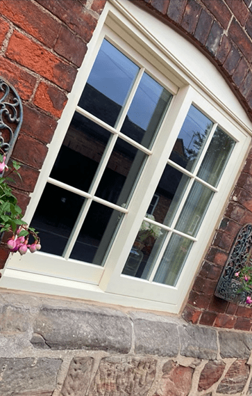 This image captures a beautifully preserved window with Georgian astragals, painted in off-white, contrasting against the rich red brickwork of an old building, complemented by decorative ironwork and blooming flowers.