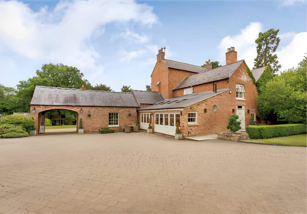 A beautifully restored traditional brick country house with a mix of pitched and flat roofs. The property features elegant timber sliding sash windows and a Churchill-style entrance door, blending heritage charm with modern craftsmanship. A large paved driveway leads up to the house, surrounded by neatly trimmed hedges and lush greenery