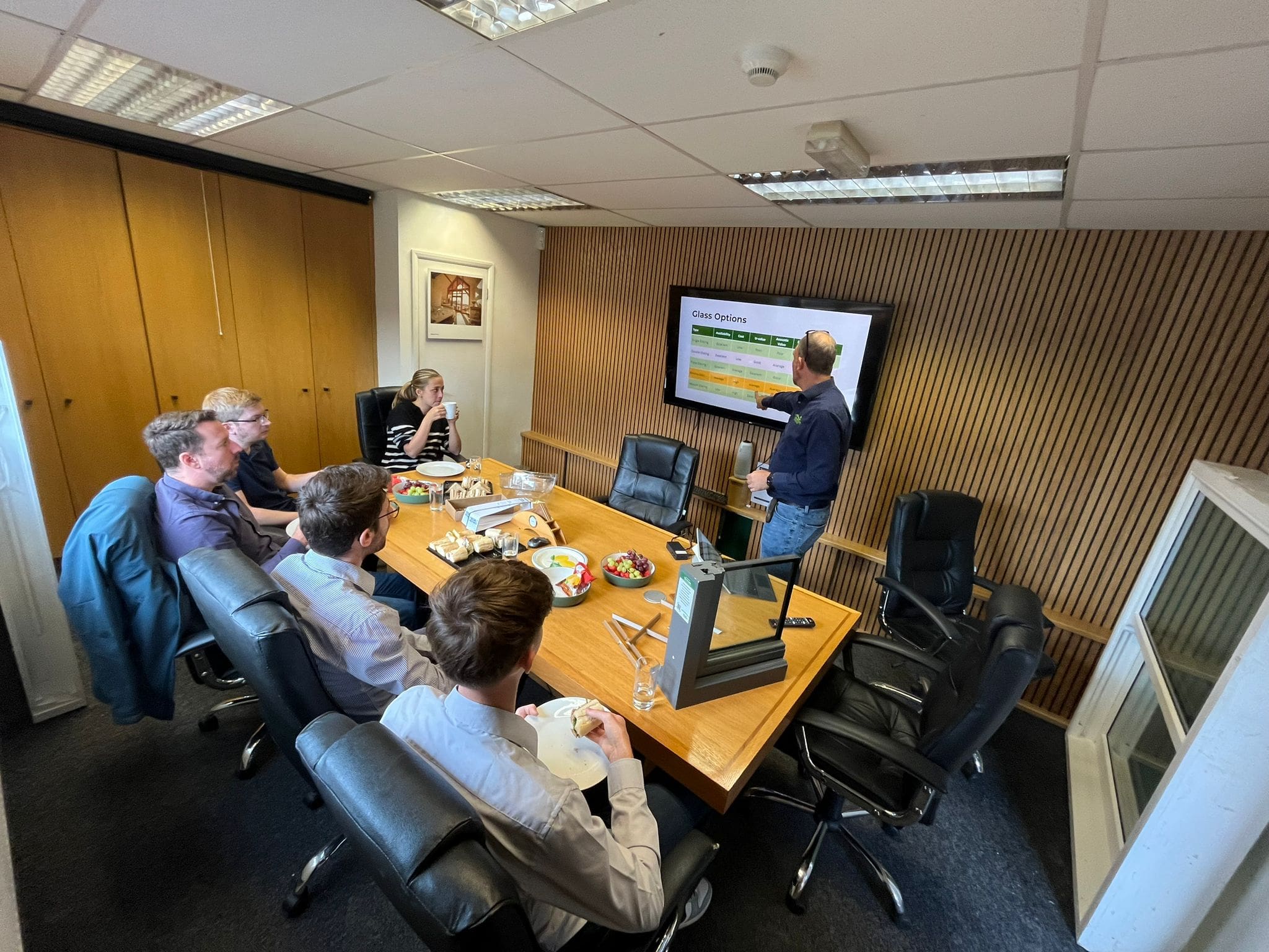 Architects attending a CPD session on windows and doors, seated around a conference table while a presenter explains glazing options on a screen. A sample timber window section is displayed on the table.