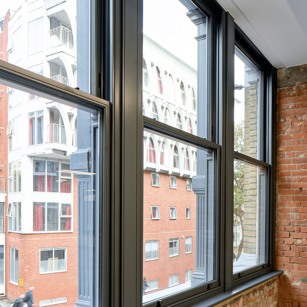 Close-up of grey-framed triple glazed Accoya Chatsworth sliding sash windows in a commercial building, set against exposed brickwork and overlooking modern city architecture.