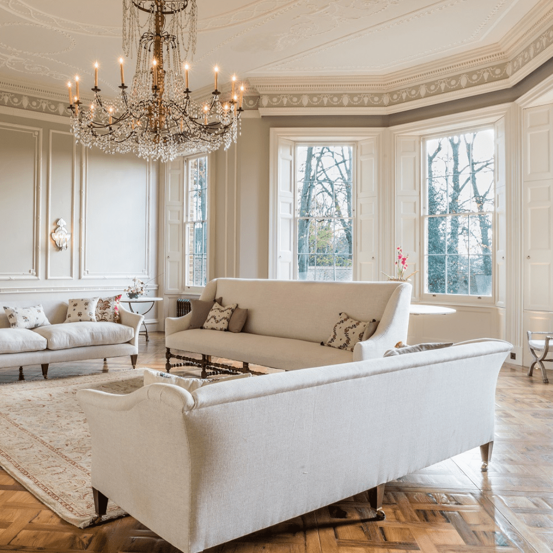 Elegant period living room with ornate plaster ceiling, chandelier, parquet flooring, and tall Winston heritage sliding sash windows framed by traditional shutters, filling the space with natural light.