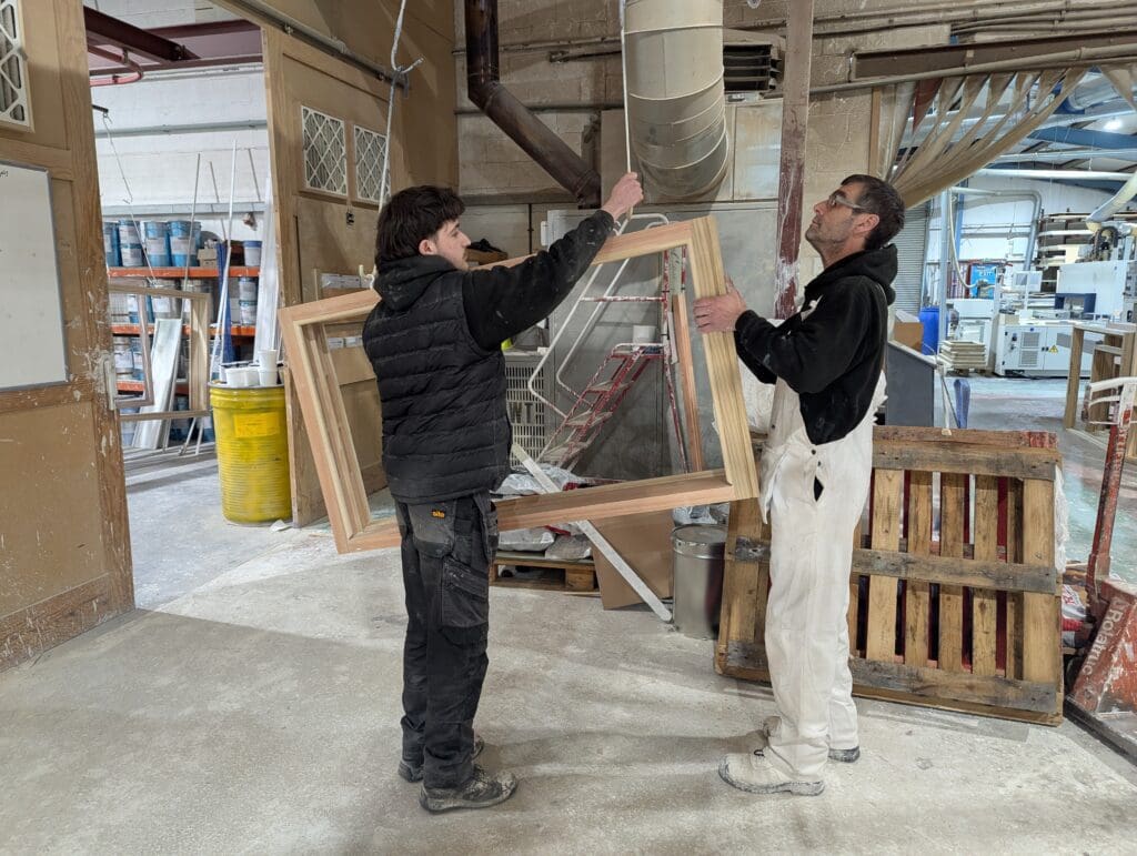 Two joiners working together in a workshop, lifting and inspecting a wooden window frame. One is wearing white overalls and safety glasses, while the other wears black workwear. Shelving, tools, and machinery are visible in the background of the busy joinery workshop.