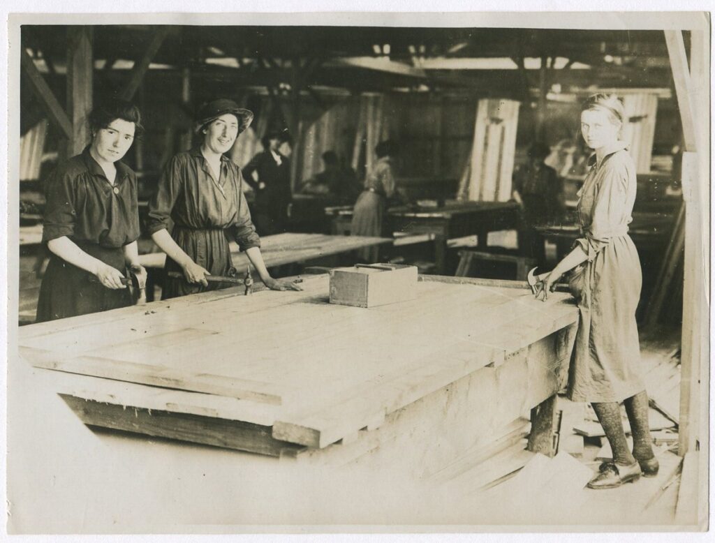 Archival black-and-white image titled "British Women Carpenters Near Front." Several women work on wooden hut panels inside a timber-framed workshop during World War I. They wear long smocks or dresses and are using hand tools at sawhorses. Stacks of timber and tools are visible in the background. These women were part of the Tarrant Hut construction effort near the front lines in France.