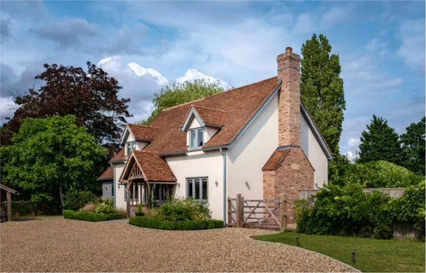 Detached country-style house with a red tiled roof, white render, brick chimney, timber porch and gravel driveway, surrounded by mature trees and garden planting.